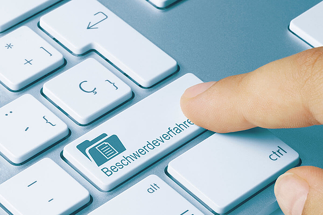 Ausschnitt einer Tastatur mit Wort Beschwerdeverfahren auf der Enter-Taste und Zeigefinger, der darauf klickt.